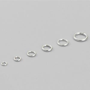 May include: Six silver jump rings of different sizes are displayed against a grey backdrop. The rings are circular and appear to be made of metal. The sizes vary, with the smallest ring on the left and the largest on the right.