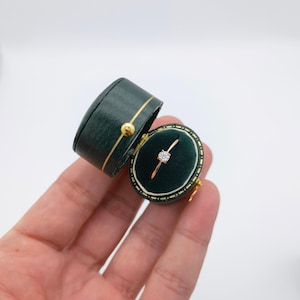 Könnte beinhalten: Eine kleine, grüne Ringbox mit goldener Einfassung ist geöffnet und zeigt einen schlichten Goldring mit einem einzelnen Diamanten. Die Box wird in einer Hand gehalten.