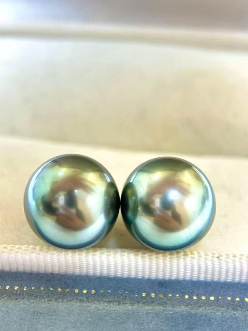 Peut inclure: Deux perles rondes, iris&eacute;es, de couleur sarcelle. Les perles ont une surface lisse et r&eacute;fl&eacute;chissante, avec des reflets dor&eacute;s et argent&eacute;s. Les perles sont pr&eacute;sent&eacute;es sur une surface bleu clair et blanche.