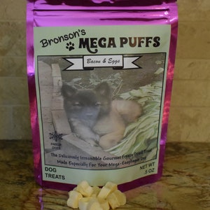 Puede incluir: Una bolsa rosa resellable con una etiqueta blanca que dice "Bronson's MEGA PUFFS Bacon & Eggs" y una foto de un cachorro. La etiqueta también dice "Freeze Dried" y "The Deliciously Irresistible Gourmet Freeze Dried Treat Made Especially For Your Mega-Esophagus Dog" y "Dog Treats" y "Net Wt 0.5 oz". Hay golosinas para perros liofilizadas en forma de corazón delante de la bolsa.