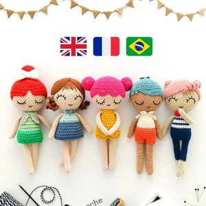 May include: Six crocheted dolls with different colored hair and clothing. The dolls are standing on a white surface with a string of flags above them. The flags are red, white, and blue, green, yellow, and blue, and green, yellow, and blue. The dolls are all wearing different outfits, including a green dress, a blue dress, a yellow dress, a red and white shirt, a blue and white shirt, and a striped shirt.