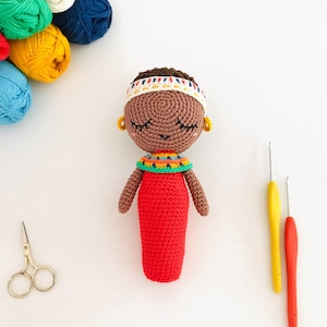 Könnte beinhalten: Eine gehäkelte Puppe mit brauner Haut, geschlossenen Augen und einem roten Kleid. Die Puppe trägt eine bunte Perlenkette und ein weißes Stirnband mit braunem Besatz.