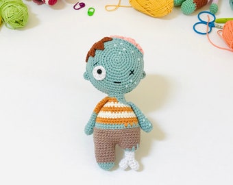 Könnte beinhalten: Eine gehäkelte Zombie-Puppe mit grünem Kopf, braunen Haaren und einem gestreiften Shirt. Die Puppe hat ein Bein und ein Knochen ragt aus dem anderen Bein heraus.