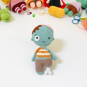 May include: A crocheted zombie doll with a green head, brown hair, and a striped shirt. The doll has one leg and a bone sticking out of the other leg.