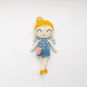 Puede incluir: Muñeca de ganchillo hecha a mano con sombrero y pelo amarillo, vestido azul y bolso rosa. La muñeca tiene los ojos cerrados, alas celestes y zapatos amarillos. El artículo está hecho de hilo.