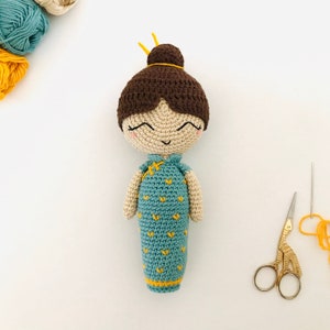 Könnte beinhalten: Eine gehäkelte Puppe mit braunen Haaren, einem blauen und gelben Kleid und einem fröhlichen Gesichtsausdruck.