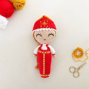 Pode incluir: Uma boneca de crochê vestindo um vestido vermelho e branco com uma borda dourada e um chapéu vermelho com uma estrela dourada. A boneca tem uma longa trança loira e está sorrindo.