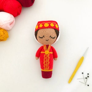 May include: A crocheted doll wearing a red and yellow dress and a white and yellow hat. The doll has brown eyes and a smiling face. The doll is sitting on a white surface with a yellow crochet hook and black pins.