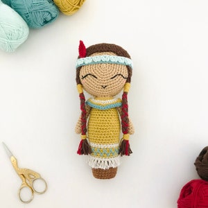 Puede incluir: Una muñeca de crochet con cabello castaño, un vestido amarillo y una diadema azul y blanca. La muñeca tiene una cara sonriente y lleva una pluma roja en la cabeza.