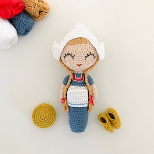 May include: A crocheted doll wearing a white bonnet and blue and white dress. The doll has blonde yarn hair and red yarn accents. The doll is sitting on a white surface.