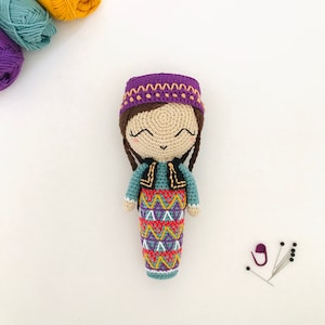 May include: A crocheted doll with brown hair, wearing a purple hat with a brown trim, a teal and brown vest, and a colorful skirt. The doll is smiling and has a happy expression.