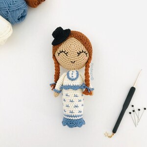 May include: A crocheted doll with brown hair, a black hat, and a white and blue dress. The doll is smiling and has closed eyes. The doll is sitting on a white surface.