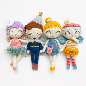 May include: Four crocheted dolls with different outfits and accessories. The dolls have closed eyes and smiling expressions. One doll is wearing a blue and gold prince costume, another is wearing a blue dress with a pink bag, another is wearing a pink dress with a flower on her head, and the last doll is wearing a yellow dress with a striped top.