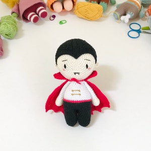 May include: A crocheted vampire doll with a red cape, white shirt, and black pants. The doll has a black and white face with a small smile and fangs.