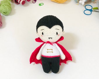 May include: A crocheted vampire doll with a red cape, white shirt, and black pants. The doll has a black and white face with a small smile and fangs.