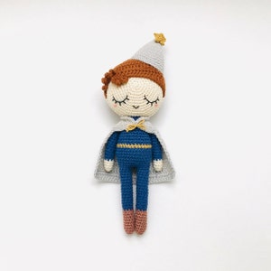 以下が含まれることがあります： 青い金色の服を着た、グレーのケープと星型の帽子をかぶった、かぎ針編みのぬいぐるみ。ぬいぐるみは目を閉じ、笑顔をしています。