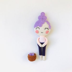 Pode incluir: Uma boneca de crochê com cabelo roxo, uma camisa listrada rosa e branca e calças azuis. A boneca está sentada ao lado de uma pequena cesta marrom com ovos roxos dentro.
