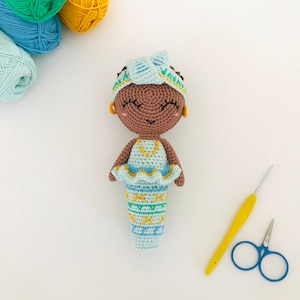 Peut inclure: Une poupée sirène en crochet avec une peau brune, une queue à motifs bleus et verts et un bandeau bleu avec une fleur jaune. La poupée sourit et a des yeux noirs.