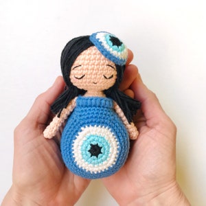 May include: A crocheted doll with black hair, a blue dress, and a blue and white evil eye design. The doll is wearing a blue and white evil eye headband.