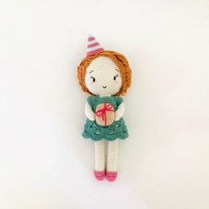 May include: A crocheted doll wearing a green dress and a pink and blue party hat. The doll is holding a small brown gift box with a red ribbon.