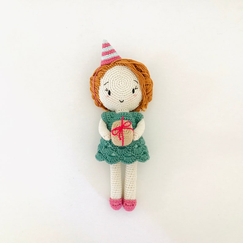 May include: A crocheted doll wearing a green dress and a pink and blue party hat. The doll is holding a small brown gift box with a red ribbon.
