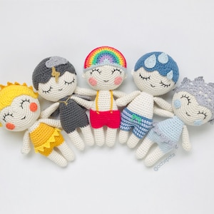 Pode incluir: Cinco bonecas de crochê com roupas e expressões faciais de diferentes cores. As bonecas usam todas roupas diferentes, incluindo uma camisa arco-íris, uma camisa listrada azul e branca, um vestido amarelo, um vestido cinza e um vestido azul. As bonecas estão todas sorrindo e têm os olhos fechados.