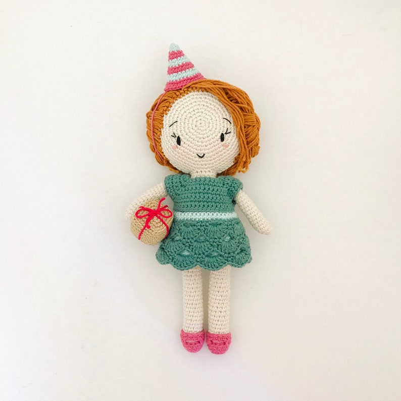 May include: A crocheted doll wearing a green dress, a party hat, and holding a small gift. The doll has brown hair and a smiling face.