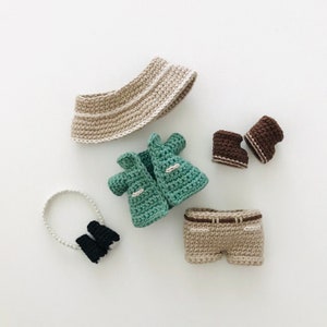 以下が含まれることがあります： 緑色のジャケット、ベージュのショートパンツ、ベージュの帽子、茶色のブーツを含む、人形用の編み物製の服のセットです。