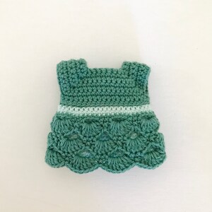 May include: A crocheted green dress with a white trim around the waist. The dress has a scalloped hem.