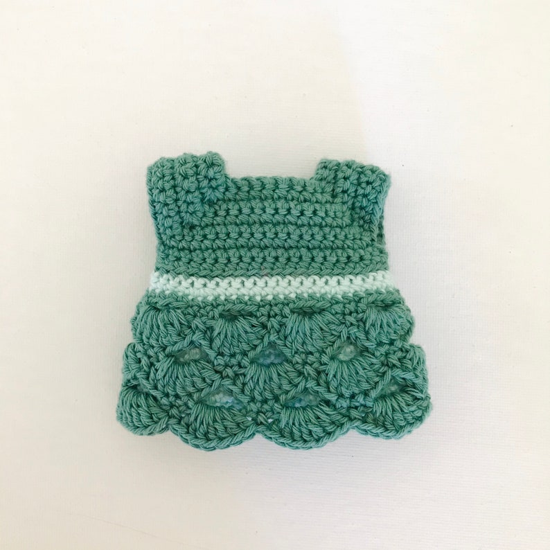May include: A crocheted green dress with a white trim around the waist. The dress has a scalloped hem.