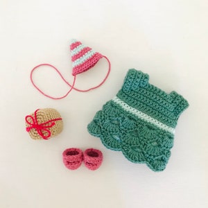 May include: A crocheted doll outfit set with a green dress, pink shoes, a pink and white striped hat, and a brown gift box with a red ribbon.