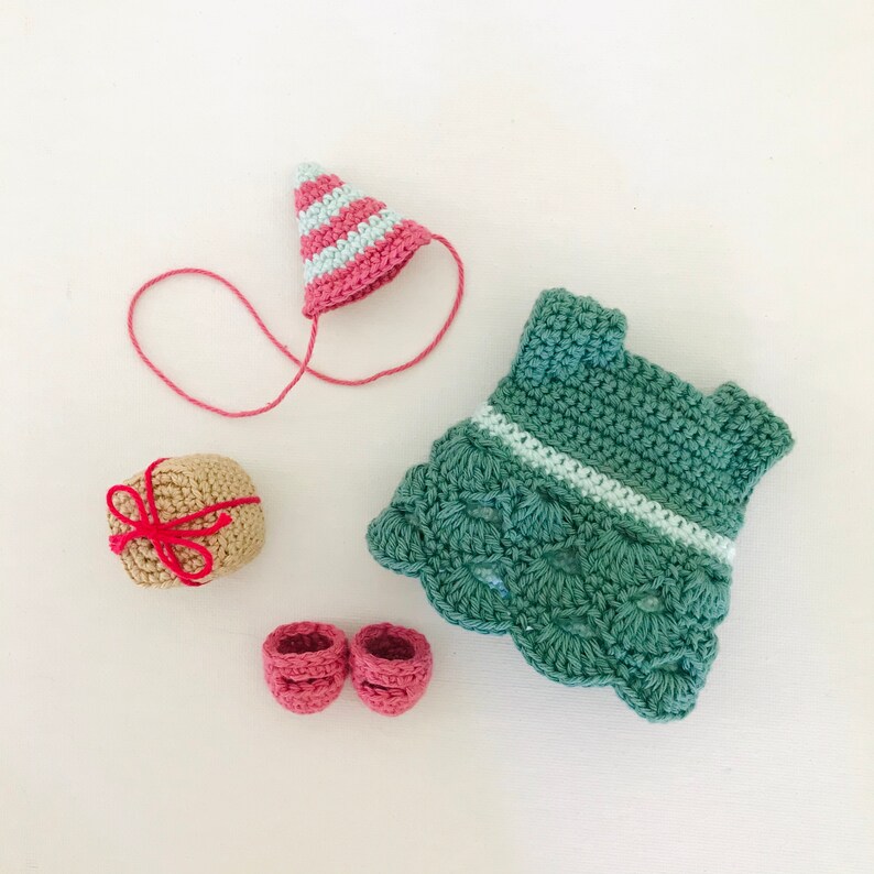 May include: A crocheted doll outfit set with a green dress, pink shoes, a pink and white striped hat, and a brown gift box with a red ribbon.