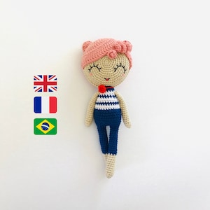 Puede incluir: Una muñeca de crochet con pelo rosa, camisa a rayas azul y blanca y pantalones azules. La muñeca tiene un lazo rojo y los ojos cerrados.