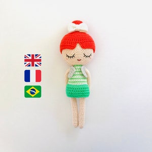 Può includere: Una bambola all'uncinetto con i capelli rossi, un fiocco bianco e un vestito a righe verde e bianco. La bambola ha gli occhi chiusi e una bocca sorridente.