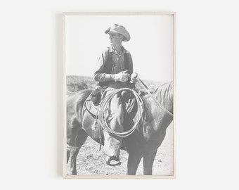 Può includere: Fotografia in bianco e nero incorniciata di un cowboy a cavallo. Il cowboy indossa un cappello, una camicia, un gilet e guanti, tenendo le redini e un lazo. Il cavallo è sellato e si trova in un campo.