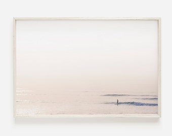 May include: A framed print featuring a serene ocean scene. The image depicts a surfer on a board in the water, with soft, muted colors of pink, white, and blue. The artwork is encased in a light-colored wooden frame.