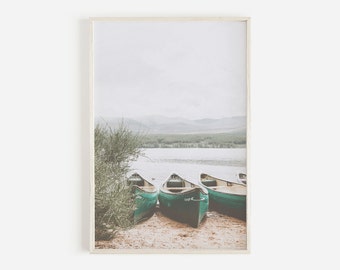 May include: A framed print featuring three green canoes resting on a sandy shore. The canoes face a calm lake with a backdrop of distant mountains and a cloudy sky. A bush is on the left side of the image.