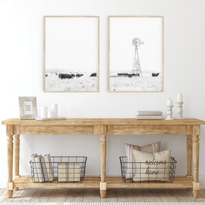 May include: A rustic wooden console table with two shelves. The top shelf features a framed print of a windmill and a framed print of a field of cows. The bottom shelf has two wire baskets, one containing books and the other with a pillow that reads "welcome home".