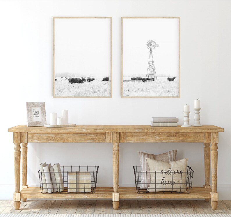 May include: A rustic wooden console table with two shelves. The top shelf features a framed print of a windmill and a framed print of a field of cows. The bottom shelf has two wire baskets, one containing books and the other with a pillow that reads "welcome home".