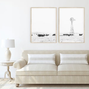 May include: Two black and white framed photographs of a rural landscape with a windmill and cattle. The photographs are hung on a white wall above a beige sofa with two white throw cushions.