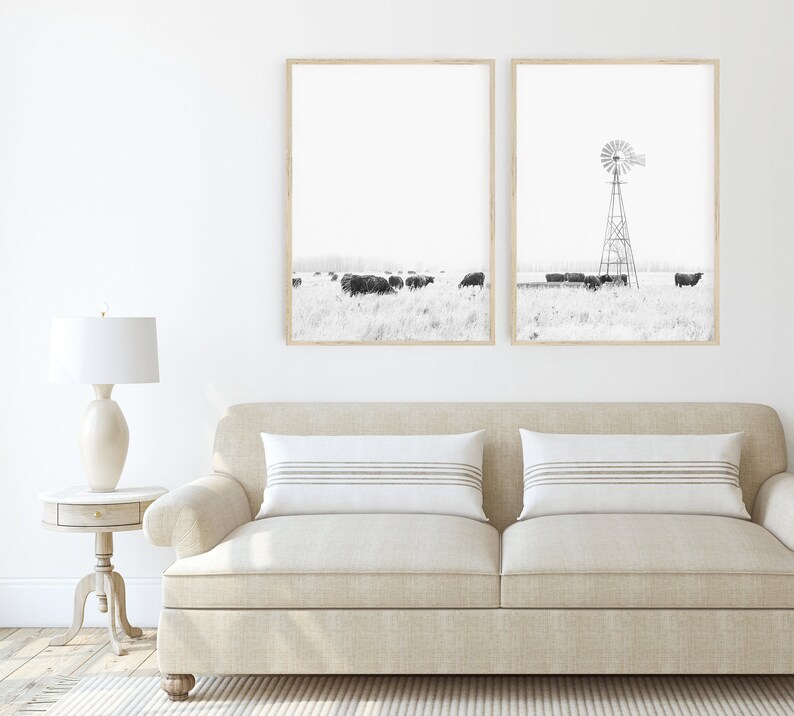 May include: Two black and white framed photographs of a rural landscape with a windmill and cattle. The photographs are hung on a white wall above a beige sofa with two white throw cushions.