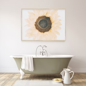May include: A framed print of a close-up of a sunflower with a light yellow and brown color scheme. The sunflower is in focus and the background is blurred. The print is hanging on a white wall above a green bathtub.