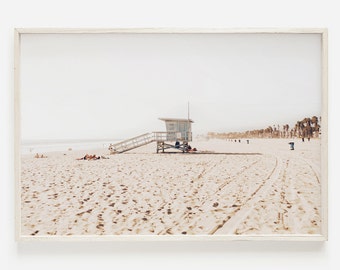 May include: A white lifeguard stand on a sandy beach with palm trees in the background. The beach is empty except for a few people sitting on the sand.