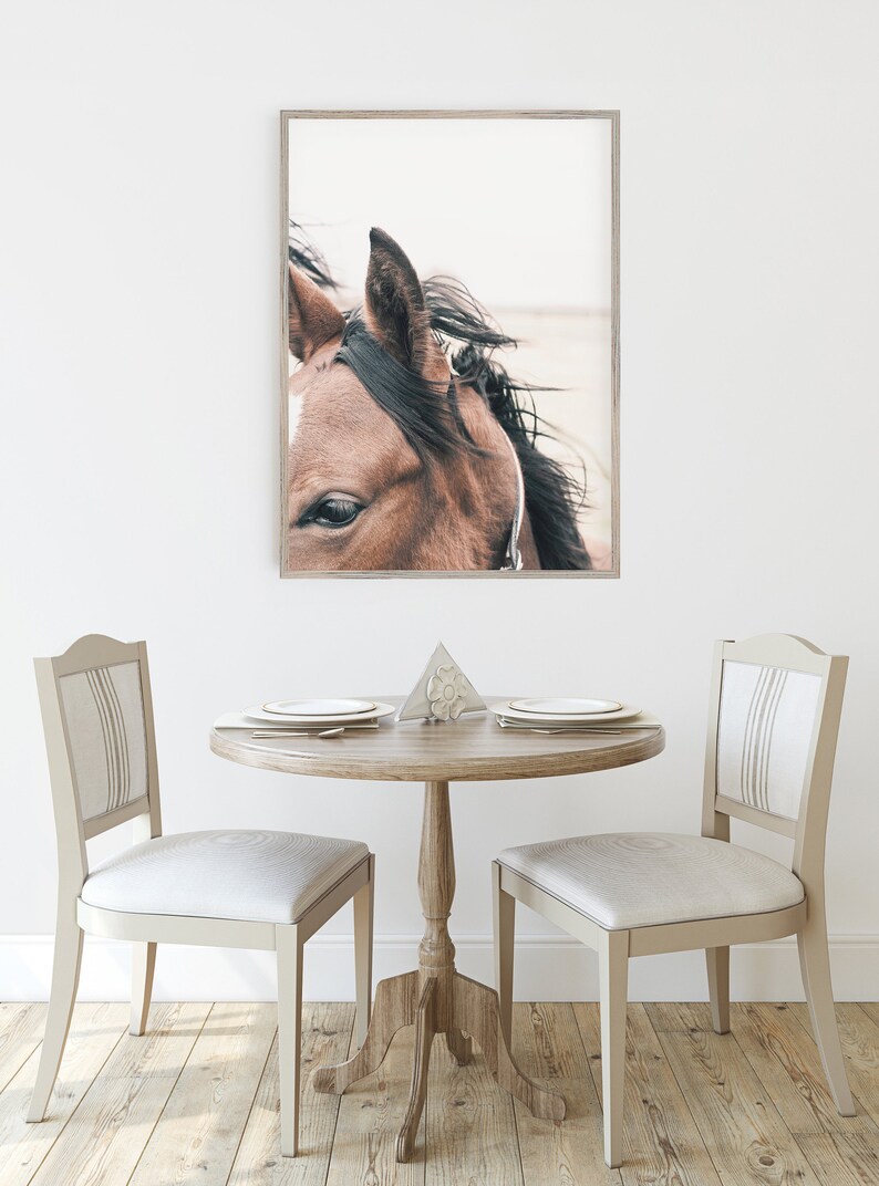 May include: A framed photograph of a brown horse's head with a white star on its forehead. The horse's mane is blowing in the wind. The photo is hanging on a white wall above a wooden table with two chairs.