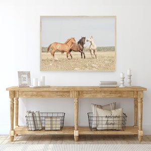 Puede incluir: Una mesa consola de madera r&uacute;stica con dos cestas de alambre, una con libros y la otra con una almohada que dice "bienvenido a casa". Una fotograf&iacute;a enmarcada de tres caballos corriendo en un campo cuelga sobre la mesa.