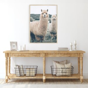 K&ouml;nnte beinhalten: Ein gerahmter Druck mit zwei Alpakas, die auf einer Wiese stehen. Die Alpakas haben hellbraunes Fell und stehen vor einem Hintergrund aus gr&uuml;nem Gras und B&auml;umen. Der Druck wird &uuml;ber einem Holztisch mit dekorativen Gegenst&auml;nden ausgestellt.