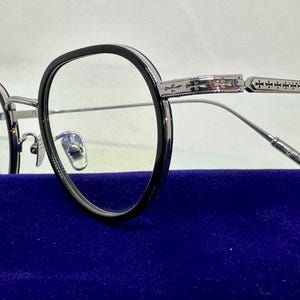 May include: Eyeglasses with black circular frames and silver-toned metal temples. The temples feature intricate engraved cross patterns along the sides. The bridge and nose pads are made of silver-toned metal, and the word TITANIUM is visible on the frame.