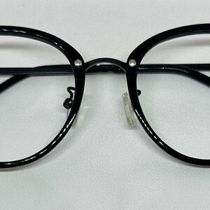 May include: Black-framed eyeglasses with a rounded shape. The glasses have a thick, glossy black frame with clear lenses. Small silver accents are visible on the outer corners of the frame. The temples are thin and black.