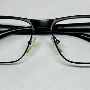 May include: Black rectangular eyeglasses with a matte finish. The glasses have a thick frame and clear lenses. The nose pads are a light yellow color. The text "VPR 61P 55016 FAD-101-140" is printed on the frame.