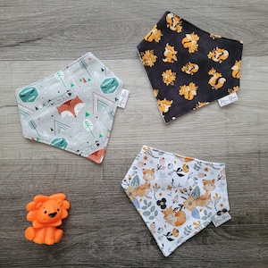 Puede incluir: Tres baberos bandana con tem&aacute;tica de zorros con diferentes patrones. Un babero es gris con acentos verdes y naranjas y el texto "Be Wild". Otro babero es negro con zorros naranjas. El tercer babero es blanco con zorros naranjas y acentos florales. Todos los baberos tienen el texto "Mini-Me" en la esquina inferior derecha.